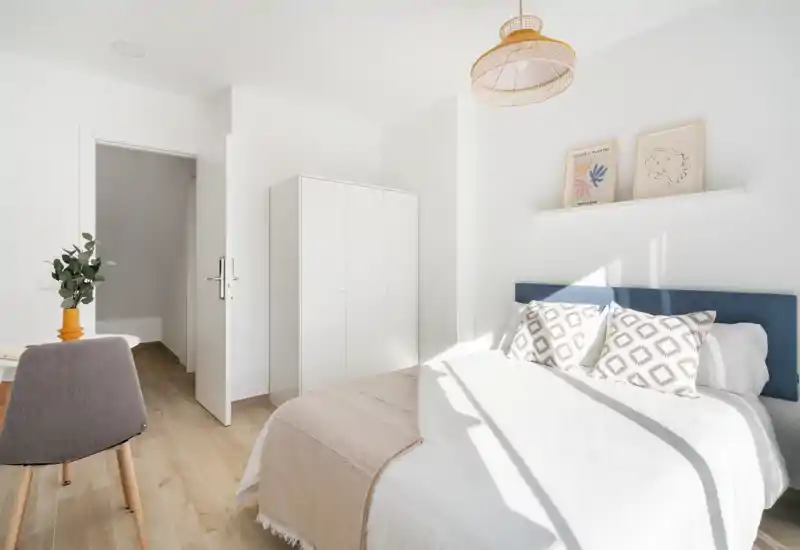 Sunny, minimalist bedroom with a double bed, patterned cushions, a wardrobe and soft natural light, ideal for rental photos.