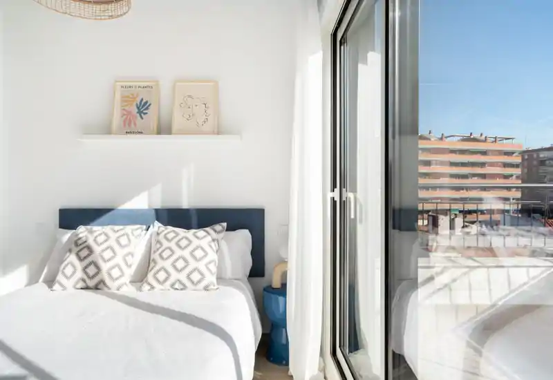 Bright, minimal bedroom with a neatly made double bed, patterned cushions, and a glass door leading to a sunny balcony — ideal for listings highlighting natural light and contemporary styling.