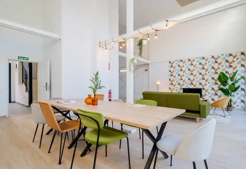Spacious, modern living area with a large communal table, green sofa, decorative patterned wall and abundant natural light — ideal for socializing or remote work.
