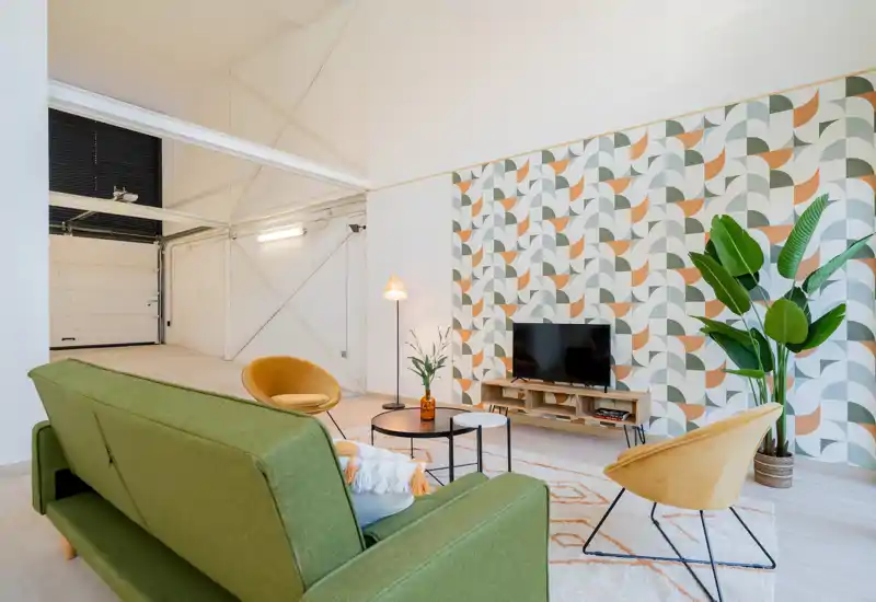 Bright, stylish living room with a green sofa, mustard accent chairs, patterned feature wall and TV — modern, cozy lounge area ideal for relaxing.