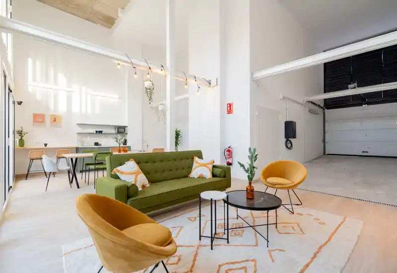 Spacious, bright living area with a green sofa, two mustard armchairs and modern nested coffee tables on a patterned rug. The open layout flows toward a dining area and small kitchen, ideal for socializing and relaxed stays.