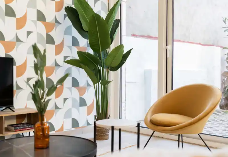 Bright, stylish living area with a comfortable mustard accent chair, round side tables and large glass doors that bring in natural light. Decorative plants and patterned wall give a modern, cozy feel ideal for relaxing.