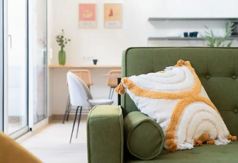Bright, modern living area with a green upholstered sofa and decorative cushion in the foreground; a small dining or workspace and sliding glass door are visible in the background.