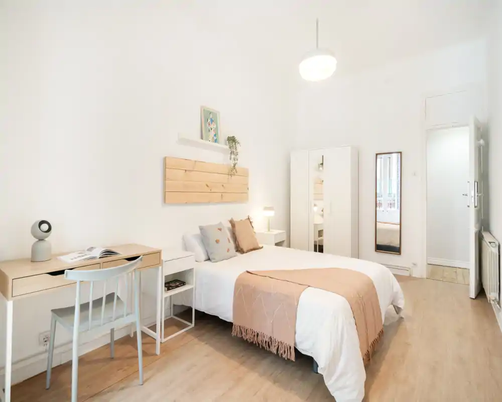Bright, minimalist bedroom with a neatly made double bed, soft throw and cushions, a small desk, wardrobe and full-length mirror — airy and well-staged for rental photos.