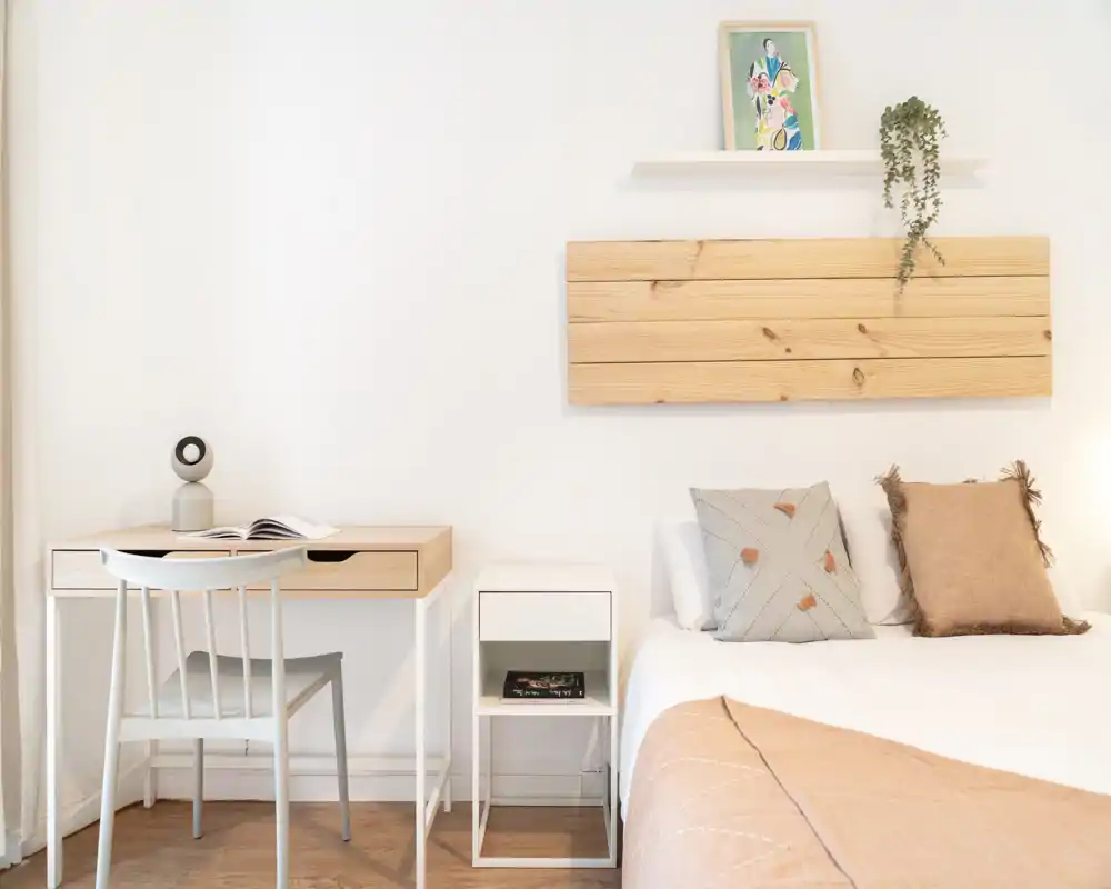 Bright, minimalist bedroom with a neatly made bed, soft throw and decorative cushions; a small desk and chair create a functional corner ideal for work or reading.