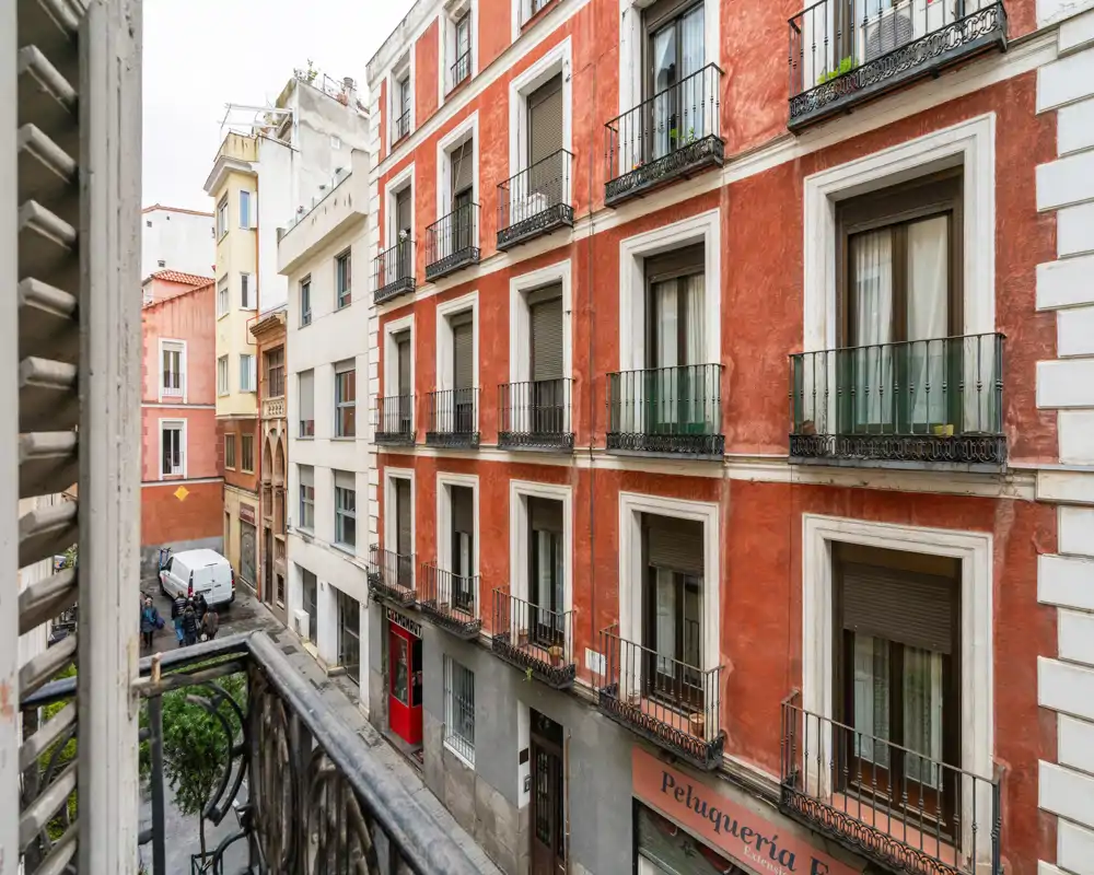 View from a small balcony overlooking a narrow street and the façade of a neighboring red building with multiple balconies.
