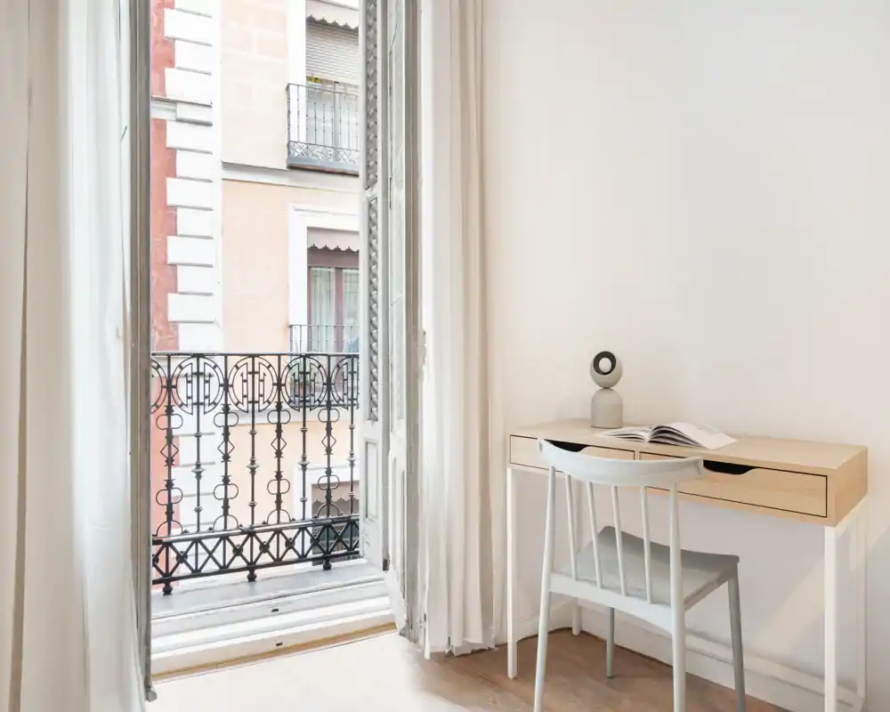 Bright, minimalist bedroom corner with an open balcony door, a simple wooden desk and chair—ideal for a calm workspace with lots of natural light.