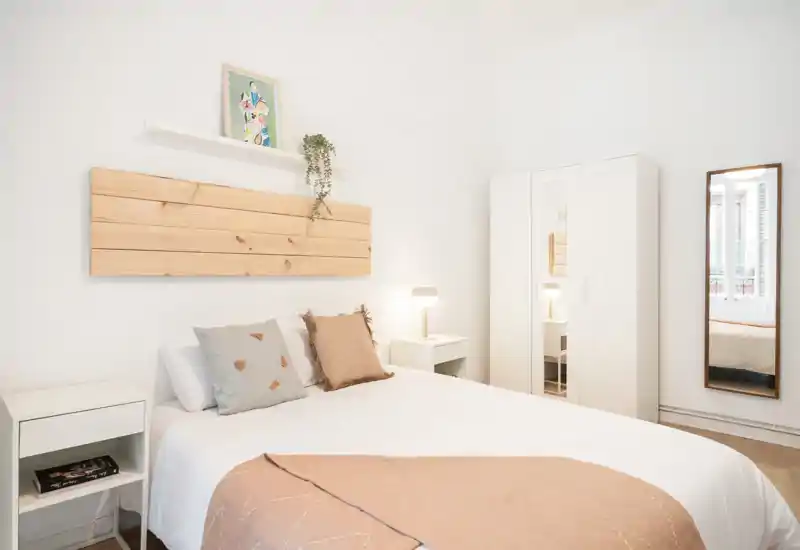 Bright, minimalist bedroom with a neatly made double bed, matching bedside tables, a wardrobe and a full-length mirror — ideal for rental listings highlighting clean, airy sleeping space.