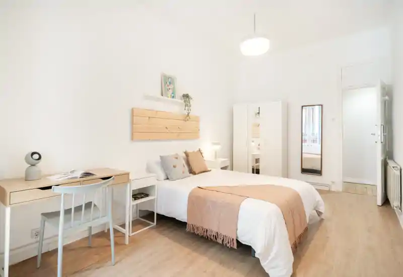 Bright, minimalist bedroom with a neatly made double bed, soft throw and cushions, a small desk, wardrobe and full-length mirror — airy and well-staged for rental photos.