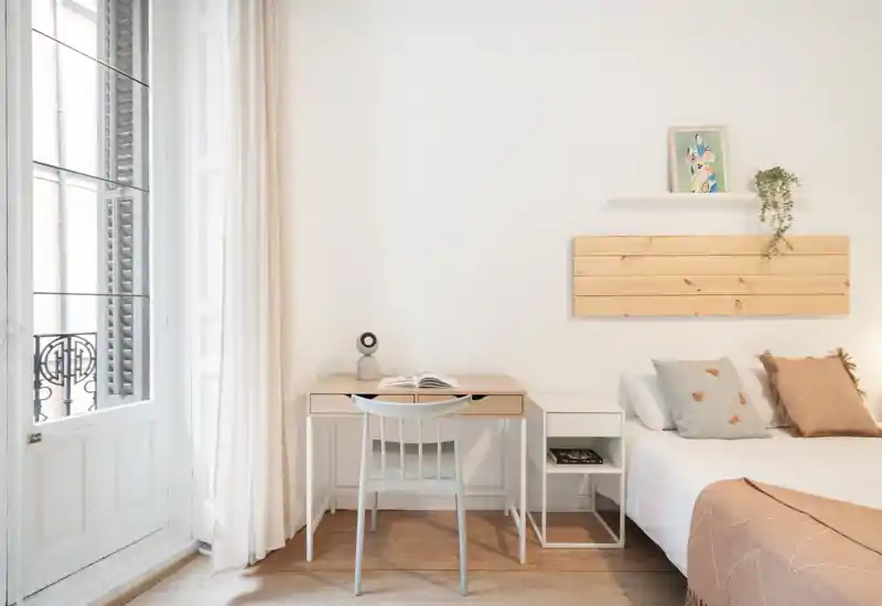 Bright, minimal bedroom with a comfortable bed, bedside table and a small desk by a large window letting in abundant natural light.