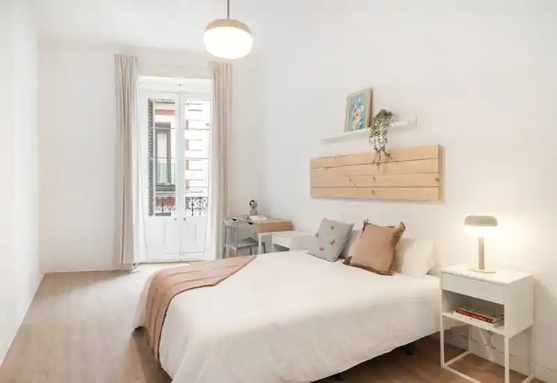 Bright, minimalist bedroom with a comfortable double bed, soft neutral textiles and a small bedside table and desk by a tall French door letting in abundant natural light.