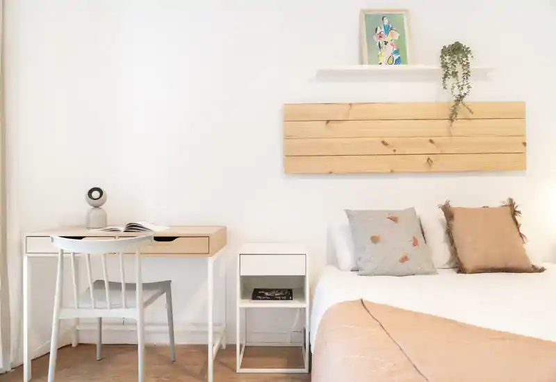 Dormitorio luminoso y minimalista con cama bien hecha, cojines decorativos y una pequeña mesita junto a un escritorio de madera clara — ideal para una estancia acogedora y funcional.