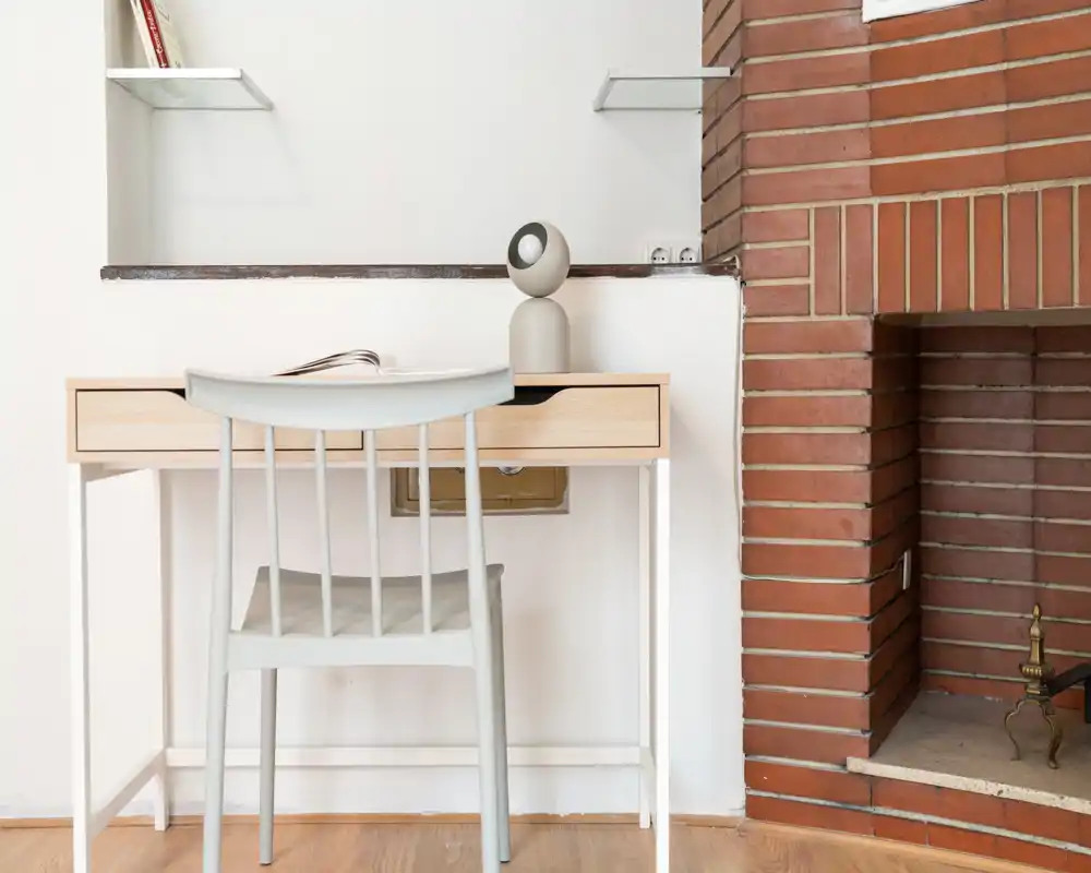 Minimal living corner with a light wooden desk, chair and small decorative lamp beside a brick fireplace — bright, tidy and styled for a cozy workspace in the living area.