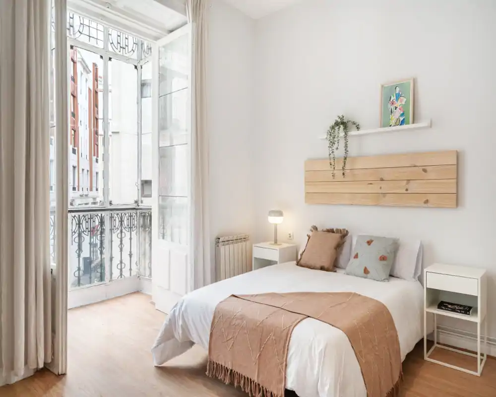 Bright, minimalist bedroom with a comfortable double bed, soft textiles and a large window opening to a small balcony — airy and well-staged for rental photos.