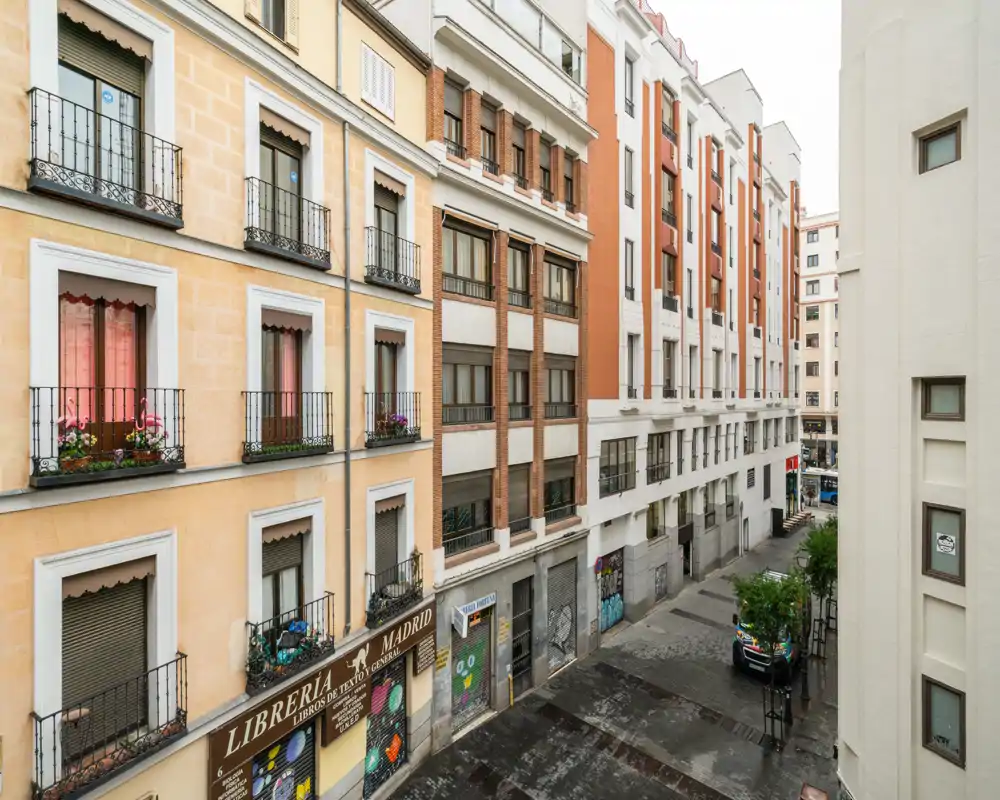 Street and neighboring building facades seen from a balcony or upper-floor window, showing a pedestrian street with storefronts and parked car.
