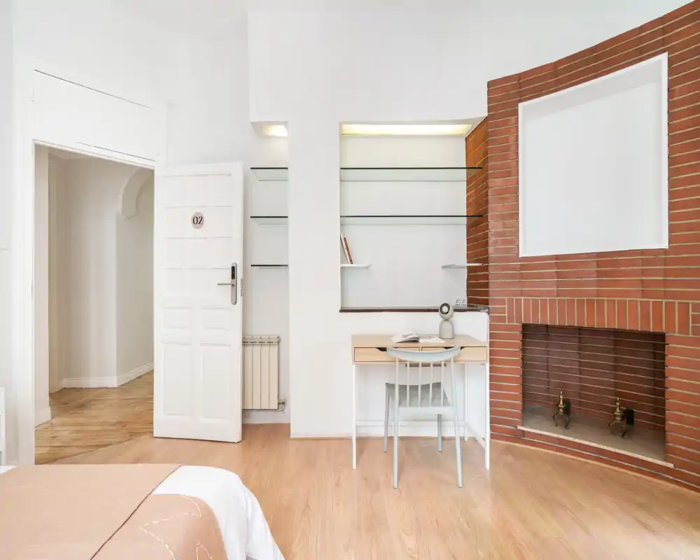 Bright, minimalist bedroom corner with a visible bed edge, built-in shelves, a small desk and a decorative brick fireplace; clean scandi-style flooring and neutral walls enhance natural light.