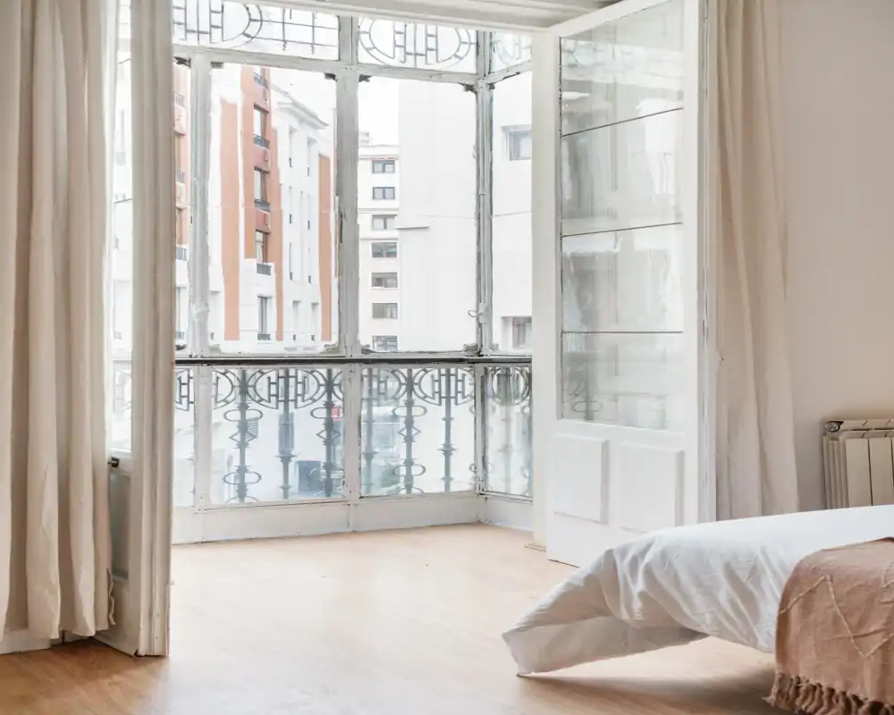 Bright, airy bedroom corner with large vintage-style windows and a partially visible bed dressed in neutral linens; light wood floors and soft curtains create a calm, inviting atmosphere.
