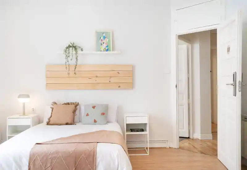 Bright, minimalist bedroom with a neatly made double bed, two white bedside tables, soft neutral textiles and warm wooden accents creating a calm, welcoming atmosphere.