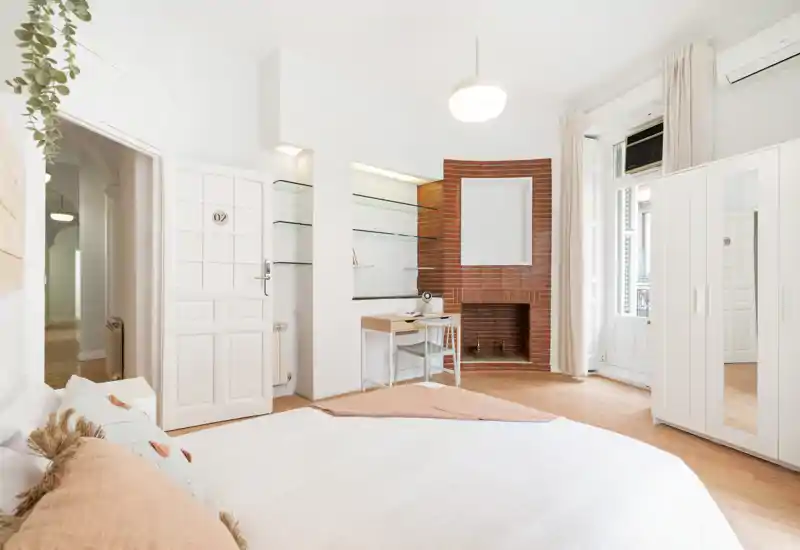 Bright, stylish bedroom with a large white bed, wooden floors, a brick decorative fireplace and a small desk by the window. Neutral linens, soft cushions and abundant natural light create a welcoming atmosphere.