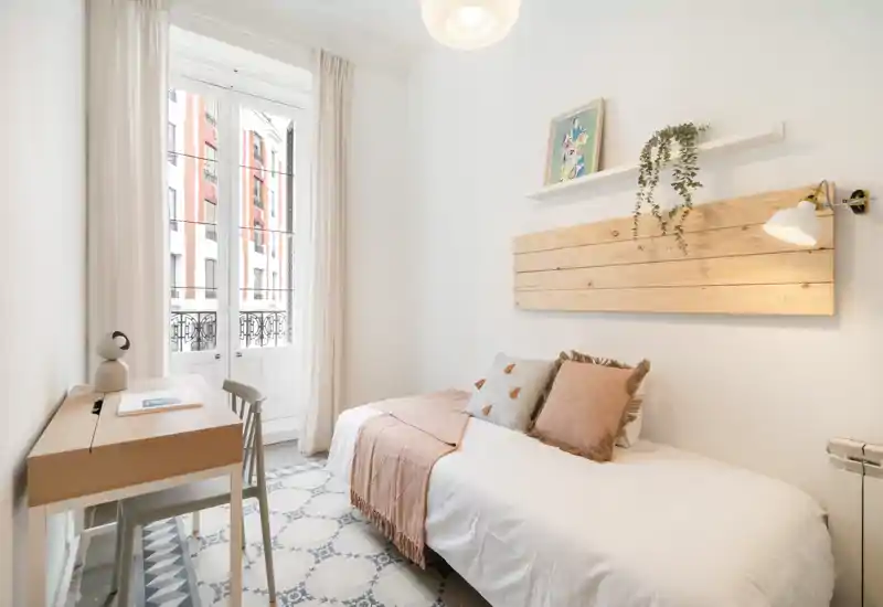 Bright, stylish single bedroom with a comfortable bed, soft textiles and a small wooden desk by a large window letting in plenty of natural light.