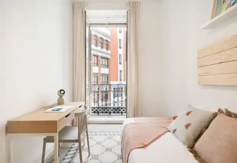 Chambre lumineuse et chaleureuse avec un lit simple confortable, des coussins moelleux et un petit bureau en bois près d'une grande fenêtre donnant sur la ville.