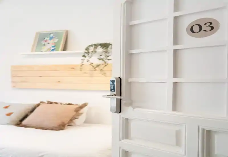 Chambre lumineuse au style doux avec linge de lit neutre, coussins décoratifs et une tête de lit en bois ; la porte entrouverte avec serrure électronique apporte une touche moderne.