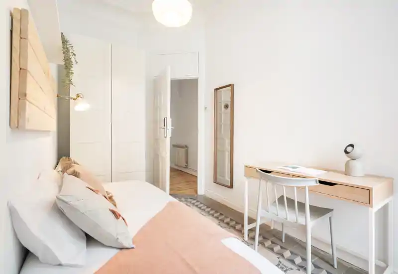 Bright, minimal bedroom with a neatly made bed, wooden desk and chair, and soft pink accents that create a calm, welcoming atmosphere.