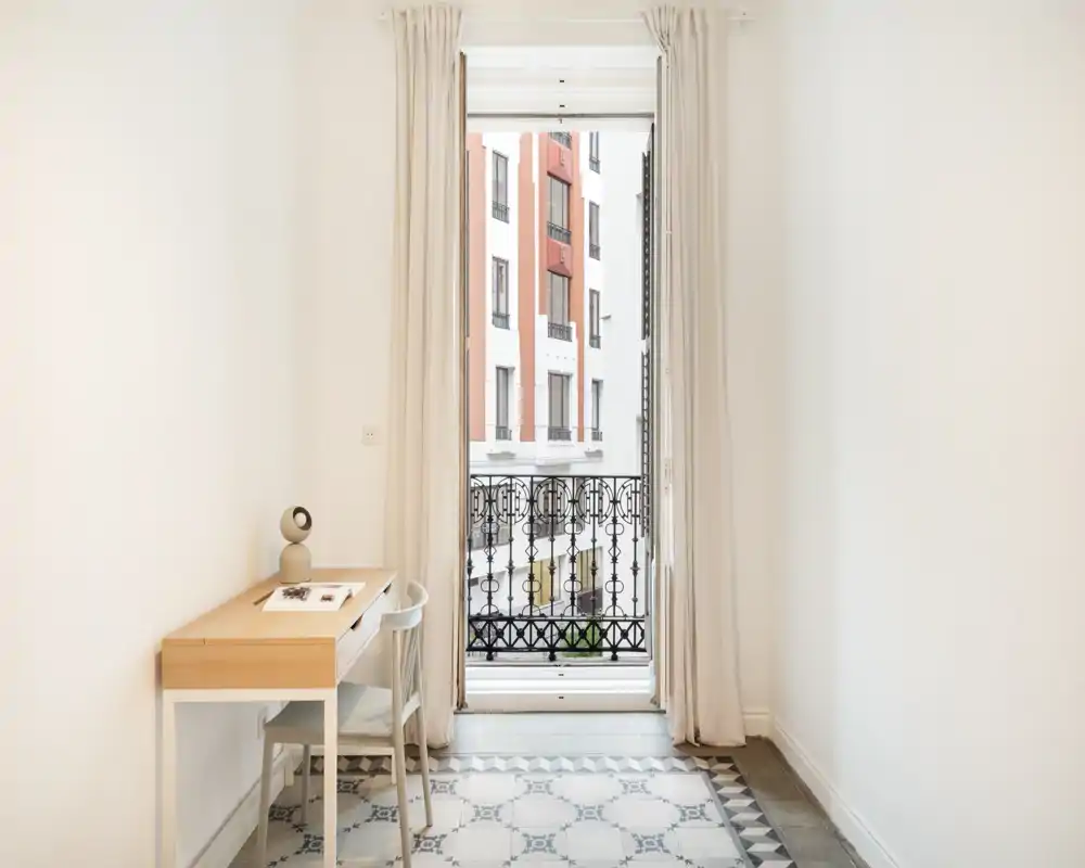 Ingresso/antiroom luminoso e stretto con una scrivania in legno, sedia e una finestra alta che si apre su un balcone con ringhiera in ferro decorata.