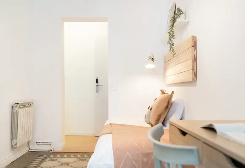 Angolo camera luminoso e minimal con un letto singolo ben rifatto, tessili caldi e una piccola scrivania—ideale per un monolocale accogliente.