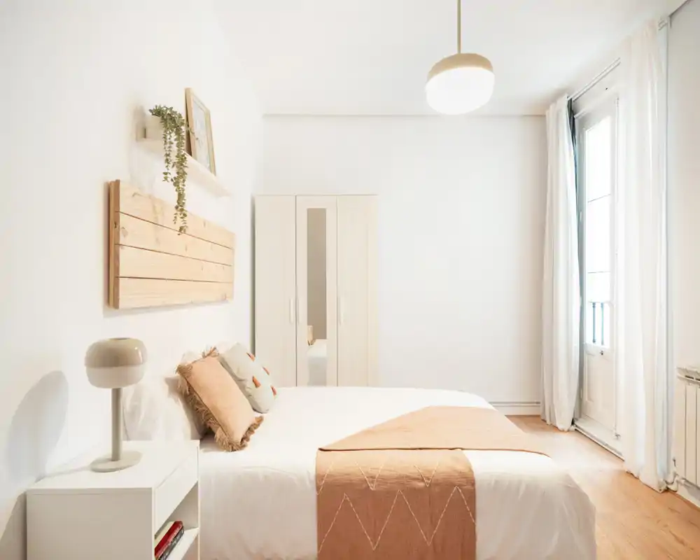 Bright, minimal bedroom with a comfortable double bed, soft cotton linens, warm accent throw and cushions, and a wardrobe next to a tall window letting in natural light.