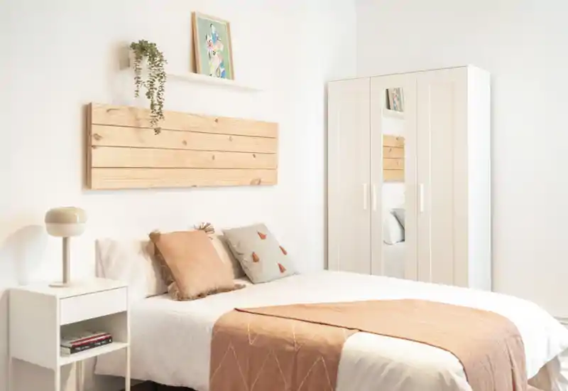 Bright, styled bedroom with a centrally framed double bed, soft linens and coordinated cushions; white wardrobe with mirrored panel and minimal Scandinavian décor.