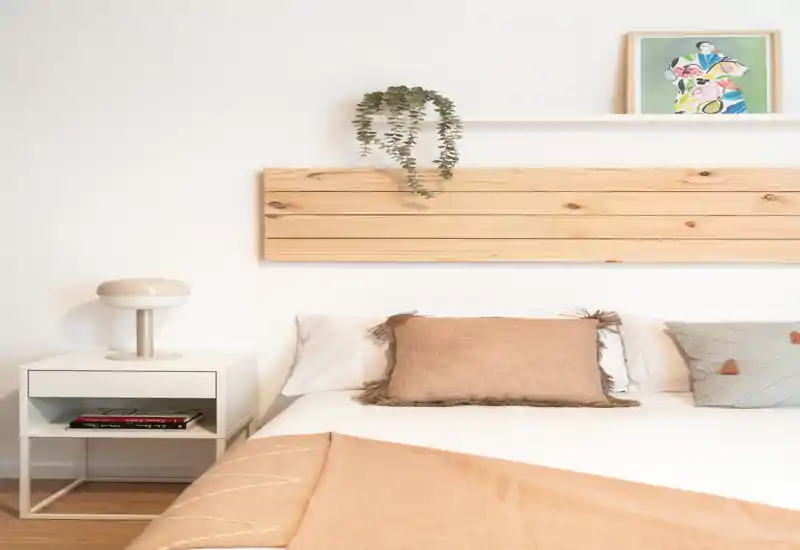 Dormitorio luminoso y minimalista con cama bien hecha, ropa de cama neutra, cojines y una mesita con lámpara; detalles de madera y una planta aportan calidez.