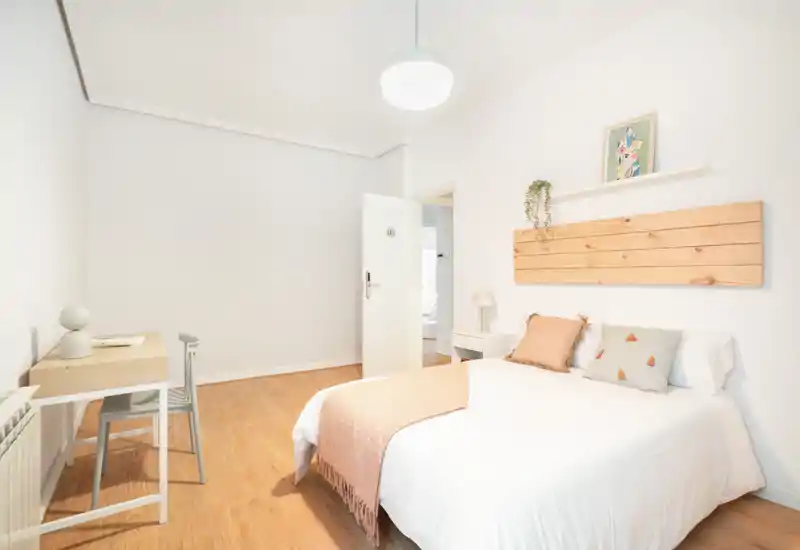 Dormitorio luminoso y minimalista con cama doble bien vestida, cabecero de madera clara y suelo de madera. Un pequeño escritorio y una silla en la pared crean un espacio para dormir y trabajar.