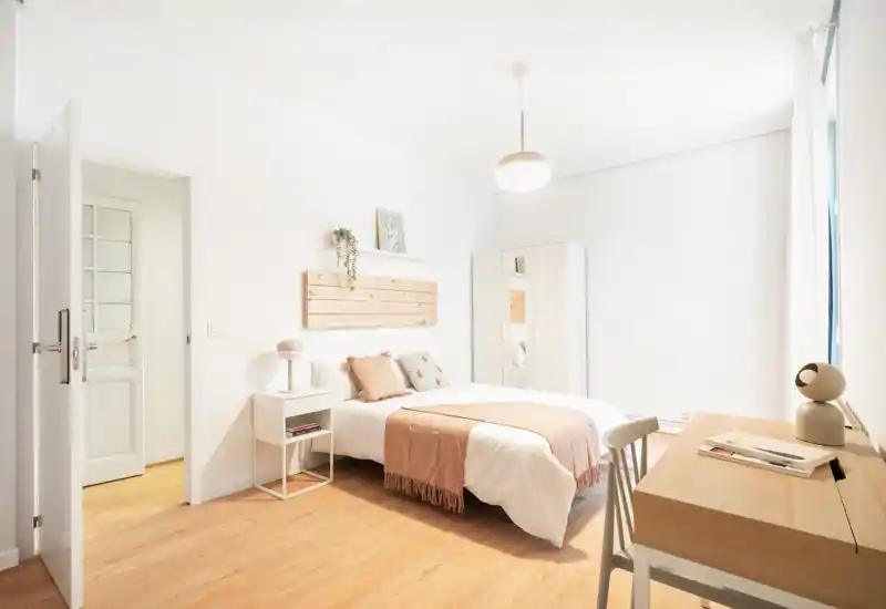Bright, modern bedroom with a neatly made double bed, warm wooden floor and simple, stylish furnishings including a desk and wardrobe.