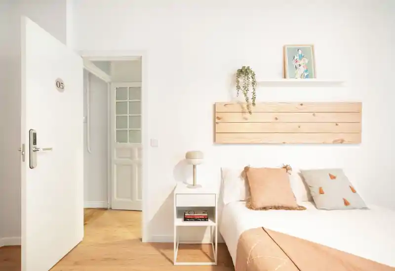 Dormitorio luminoso y minimalista con cama bien hecha, cojines suaves, mesita de noche y detalles de madera cálida: acogedor y de estilo sencillo para una estancia relajante.