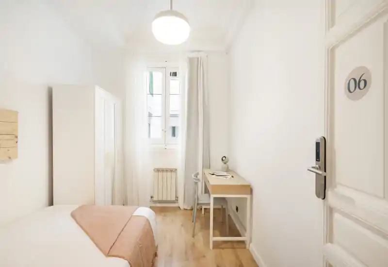 Bright, minimalist single bedroom with a neatly made bed, wardrobe, and a small desk by the window providing natural light.