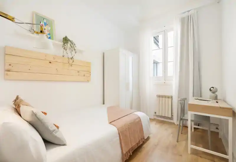 Bright, minimal bedroom with a neatly made double bed, soft throw and pillows, a wardrobe and a small desk by the window — perfect for a comfortable stay.