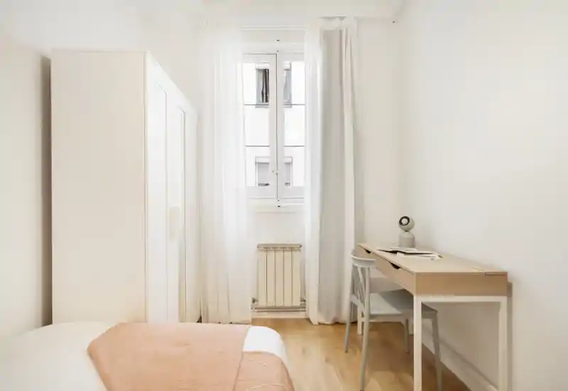 Minimal, bright bedroom with a neatly made bed, light wood flooring, a wardrobe and a compact desk by the window letting in abundant natural light.