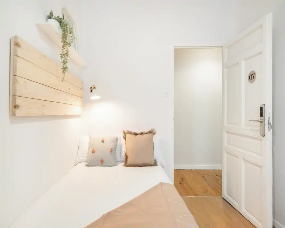 Petite chambre lumineuse avec un lit simple bien fait, des coussins moelleux et des accents en bois chaleureux. La pièce est minimaliste et accueillante, avec des murs blancs et une belle lumière naturelle.