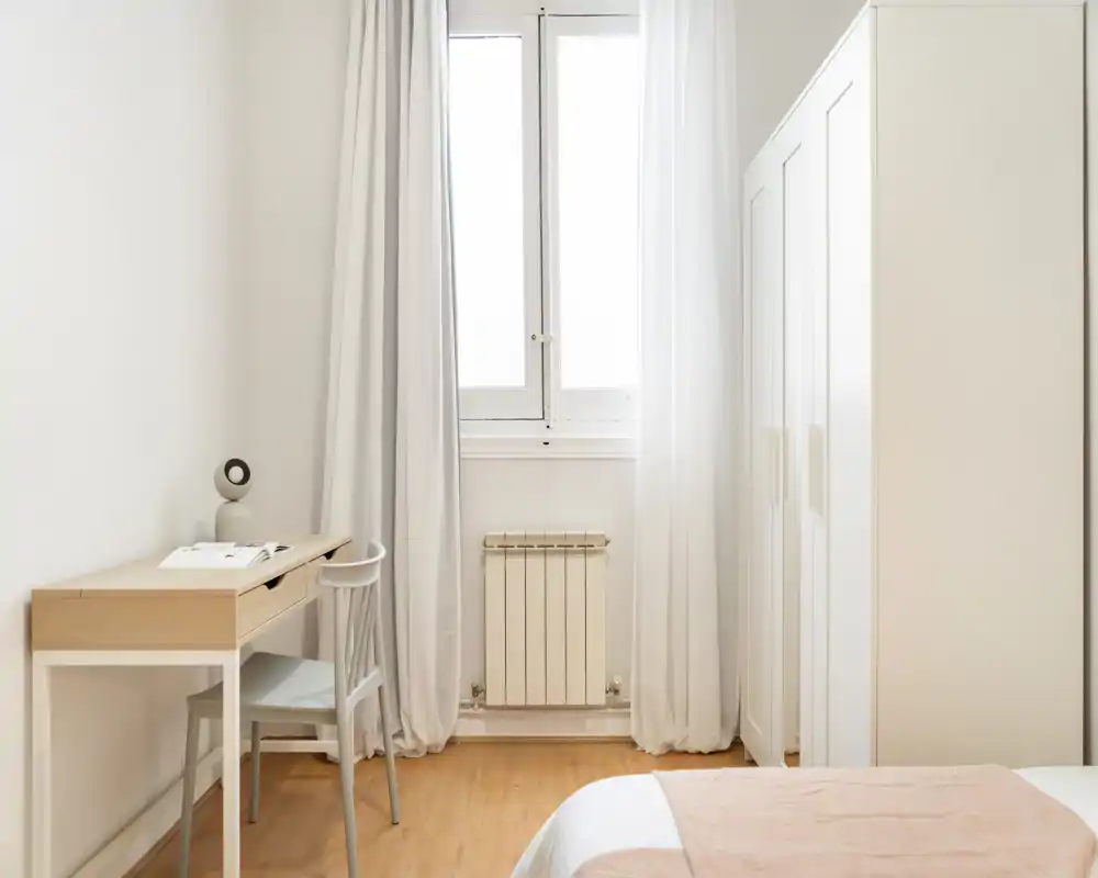 Coin chambre lumineux et minimaliste avec un lit partiellement visible, une armoire blanche et un petit bureau en bois près de la fenêtre.