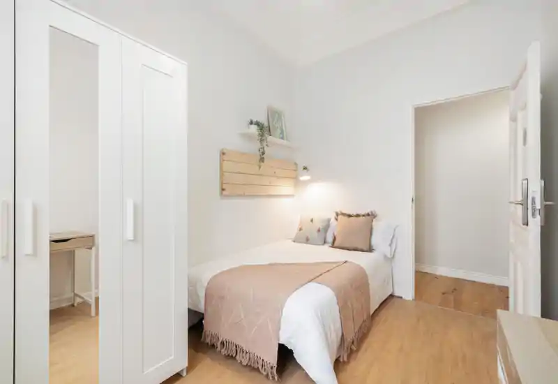 Bright, cozy bedroom with a neatly made double bed, soft beige throw and cushions, wardrobe with mirrored panel and a small desk visible — ideal for a quiet stay.
