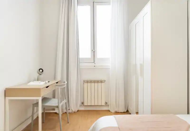 Dormitorio luminoso y minimalista con armario blanco, escritorio y cama bien hecha en tonos neutros; gran ventana que aporta abundante luz natural.