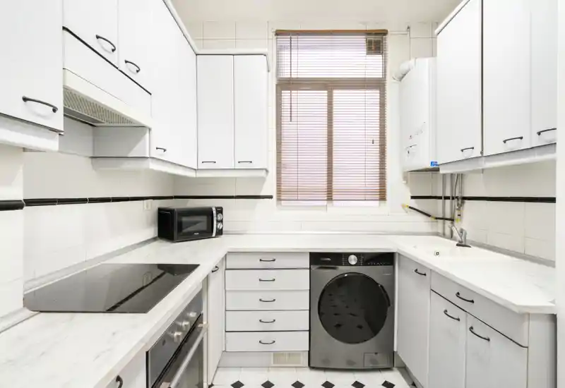 Cocina compacta y luminosa con muebles blancos, encimeras efecto mármol, placa de inducción, microondas y lavadora integrada bajo la encimera. La ventana con persiana aporta luz natural y el suelo en blanco y negro añade carácter.