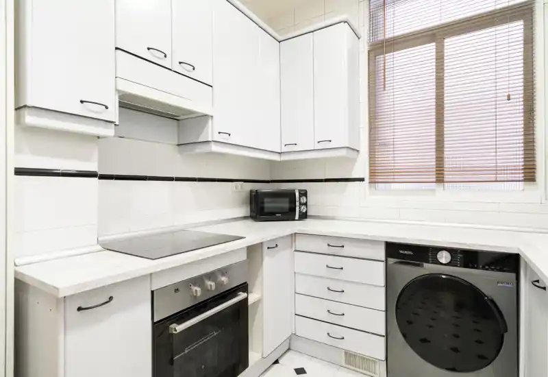 Cuisine compacte et lumineuse avec meubles blancs, plaque électrique, four et lave-linge ; plans de travail propres et une fenêtre avec stores apportant de la lumière naturelle.