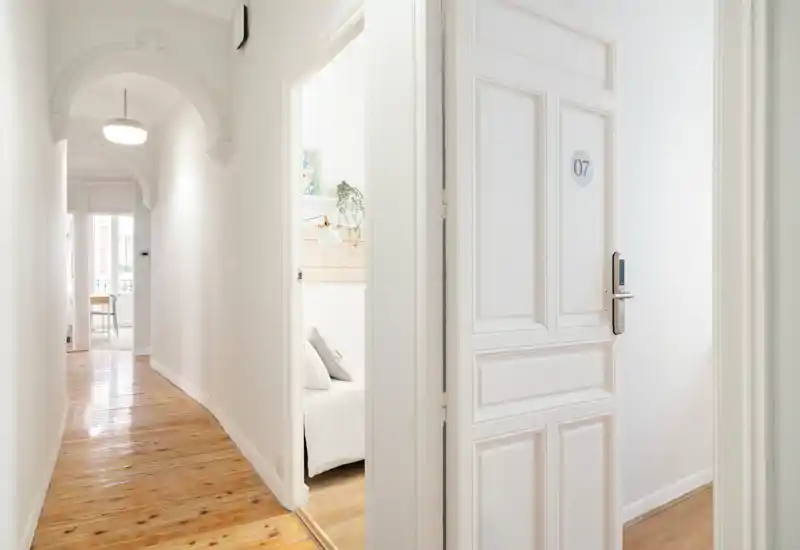 Couloir lumineux avec porte ouverte donnant sur une petite chambre sobre, linge de lit blanc et lumière naturelle douce.