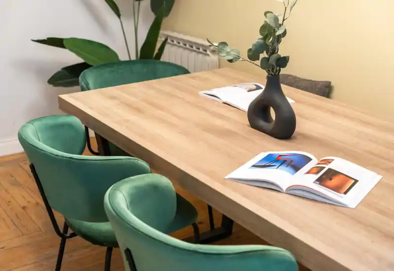 Acogedor comedor con amplia mesa de madera, sillas tapizadas verdes y un jarrón moderno; espacio luminoso y bien decorado, perfecto para comer o trabajar.