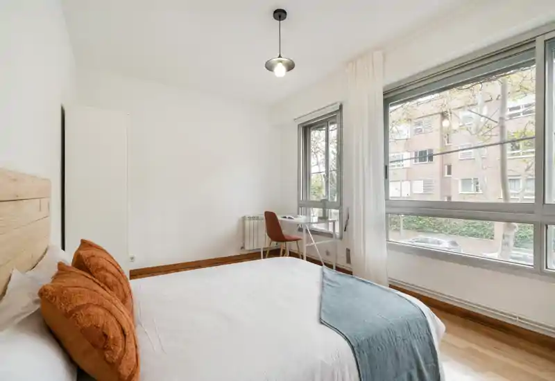 Bright, minimalist bedroom with a large window, comfortable double bed with warm cushions and a small desk by the window — ideal for restful stays and remote work.