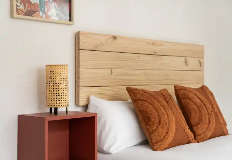 Bright, styled bedroom corner with a wooden headboard, textured rust pillows and a woven bedside lamp on a terracotta nightstand — cozy and well composed for listing photos.