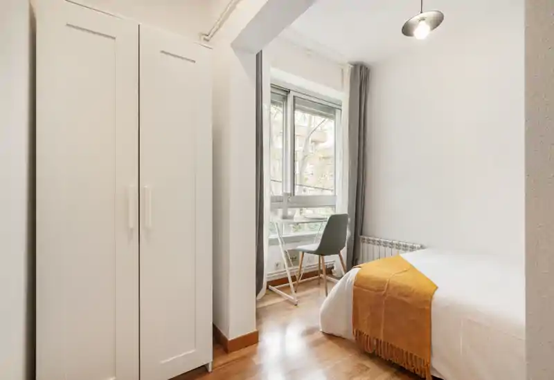 Chambre lumineuse et compacte avec un lit simple en linge blanc et un plaid ocre; une armoire et un petit bureau près de la fenêtre offrent un espace fonctionnel et minimaliste avec beaucoup de lumière naturelle.