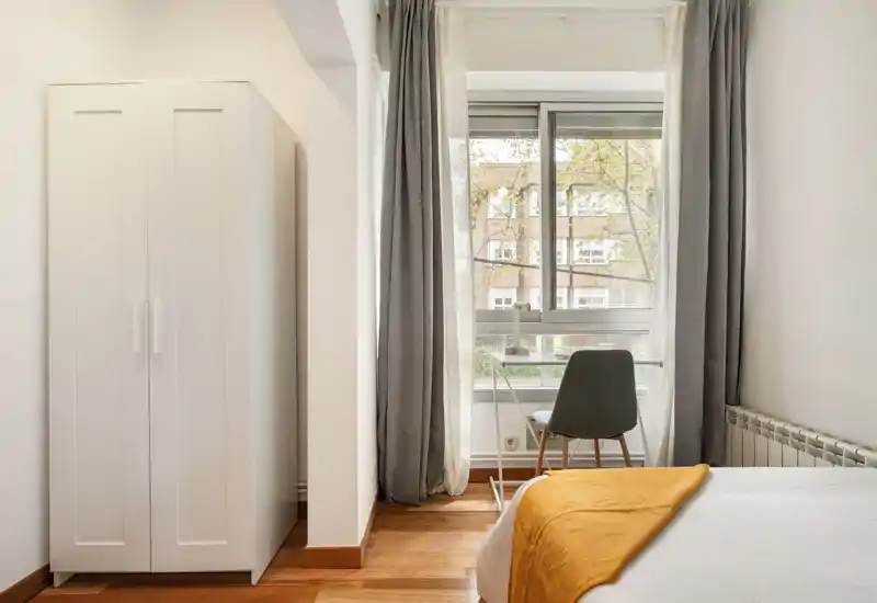 Chambre lumineuse et soignée avec un lit visible recouvert d’un jeté moutarde, une armoire et un coin bureau près de la fenêtre ; belle lumière naturelle et décoration minimaliste.